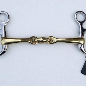Puede incluir: Un bocado para caballo con una embocadura de metal dorado con un eslabón central y piezas laterales de metal plateado con anillas. Una pequeña etiqueta rectangular negra con el texto blanco GallopGrip está sujeta a la anilla derecha.