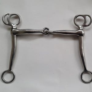 May include: A silver-coloured metal horse bit featuring a jointed mouthpiece connected to two curved shanks. Each shank terminates in a circular ring at the bottom, while the top of each shank includes a specialised ring attachment for headstall gear.
