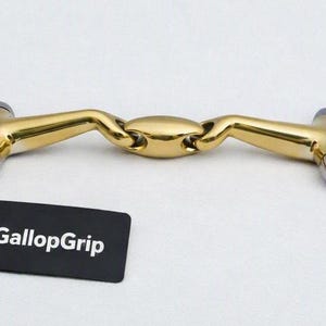 May include: A horse bit featuring a gold-colored central lozenge mouthpiece connected to two silver-colored stainless steel rings. A black rectangular tag with the white text GallopGrip is attached to one of the rings by a thin black cord.