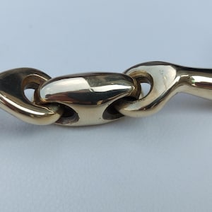 May include: A gold-coloured metal horse bit featuring a central oval-shaped link connected to two smooth, curved side bars. The polished brass-toned finish reflects light, and a silver-coloured metal ring is visible on the far right side of the bit.