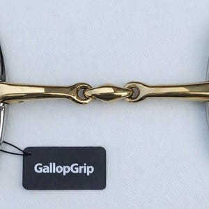 May include: A horse bit featuring a gold-coloured metal mouthpiece with a central joint and silver-coloured metal cheekpieces with multiple rings. A black rectangular tag with the white text GallopGrip is attached to the left side of the bit.