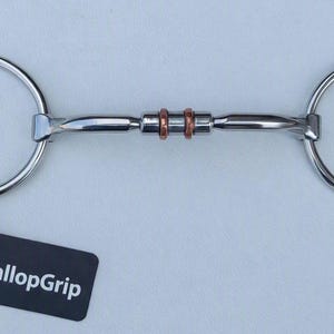 May include: A stainless steel horse bit featuring two large circular rings connected by a central bar with two copper-coloured rings in the middle. A black rectangular tag with the white text GallopGrip is attached by a thin black string.