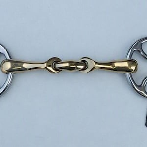 May include: A horse bit featuring a gold-colored metal mouthpiece with a central joint and two silver-colored circular cheekpieces with cut-out details. A black rectangular tag with the white text GallopGrip is attached to the right side.