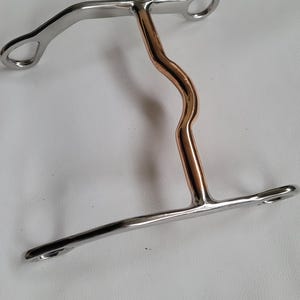 May include: A horse bit featuring a silver-colored metal frame with two circular rings at the top and a horizontal bar at the bottom. The central vertical component is made of a smooth, copper-colored metal with a distinct S-shaped curve.