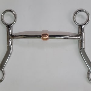May include: A silver-coloured metal horse bit featuring a curved mouthpiece with a central copper-toned roller. The bit includes two upper rings for cheek pieces and two lower rings for reins, connected by straight shanks on both sides.