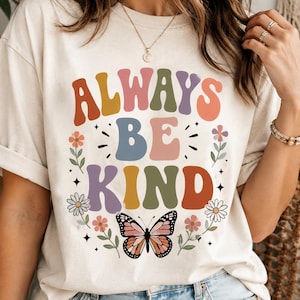 May include: A cream-colored t-shirt featuring the text ALWAYS BE KIND in multi-colored, wavy, bold lettering. The design includes a central butterfly graphic at the bottom, surrounded by small floral illustrations and star accents in shades of pink, orange, blue, and green.