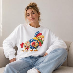 May include: A white long-sleeve sweatshirt featuring a colourful graphic of a chicken. The chicken is designed with a patchwork-style pattern in shades of blue, red, yellow, and green, decorated with floral and polka dot motifs, pecking at seeds.