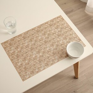 May include: A rectangular placemat featuring a repeating geometric pattern of tan and beige triangles. The placemat is positioned on a white surface alongside a clear glass filled with water and a small, plain white ceramic bowl.