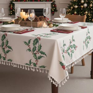 May include: A cream-coloured tablecloth featuring a repeating pattern of green holly leaves and red berries, finished with a decorative white tassel fringe along the edges. The fabric is draped over a dining table set with white plates and glassware.