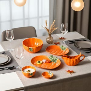 Op de afbeelding: Een set oranje keramieken servies in de vorm van pompoenen, bestaande uit een grote kom, twee ovale schalen, een sauskom, een kleine kom en een klein schaaltje. Elk stuk is voorzien van reliëf met groene pompoenbladeren en ranken.