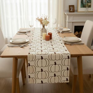 May include: A cream-colored table runner featuring a repeating pattern of dark brown outlined pumpkins. The runner is laid across a wooden dining table set with white plates, clear glassware, two pillar candles, and a vase of dried flowers.