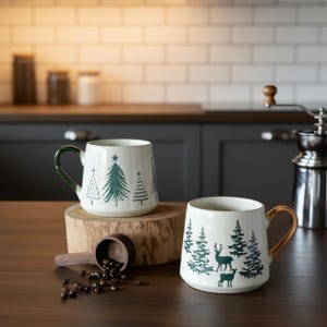 Puede incluir: Dos tazas de cerámica con base blanca e ilustraciones de bosque en verde. Una taza presenta pinos y un asa verde oscuro, mientras que la otra muestra ciervos y pinos con un asa marrón. Una cuchara de madera con granos de café está cerca.