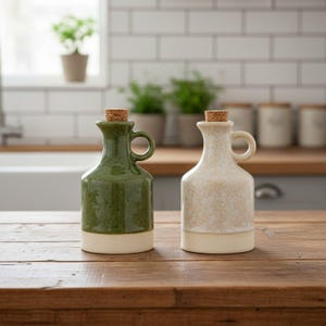 Puede incluir: Dos aceiteras de cerámica con tapones de corcho, una en verde oscuro brillante y otra en acabado crema moteado. Cada botella presenta un cuerpo redondeado, cuello corto y una pequeña asa lateral, con una base de color crema sólido.