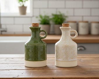 May include: Two ceramic oil cruets with cork stoppers, one in a glossy deep green and the other in a speckled cream finish. Each bottle features a rounded body, a short neck, and a small side handle, with a solid cream-colored base band.