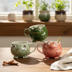 May include: Three ceramic mugs shaped like frogs in green, light green, and pink. Each mug features a rounded body, protruding eyes, and small feet at the base. A gold-toned spoon rests inside the pink mug, and two cinnamon sticks lie nearby.