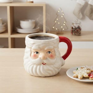 May include: A ceramic mug shaped like the face of Santa Claus with a white beard, rosy cheeks, and a bright red handle. The mug is filled with a dark liquid and is positioned next to a small white plate containing several snowflake-shaped cookies.