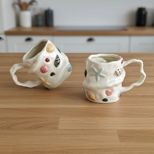 May include: Two white ceramic mugs with irregular, wavy surfaces and handles. The mugs are decorated with various small, colourful, raised seashell and starfish motifs in shades of pink, blue, yellow, and brown, scattered across the exterior.