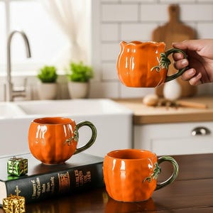 Peut inclure: Trois tasses en céramique orange en forme de citrouille avec des textures côtelées et des anses vertes ornées de détails sculptés de vignes et de feuilles. Les tasses ont une finition brillante et un corps arrondi imitant une petite citrouille.