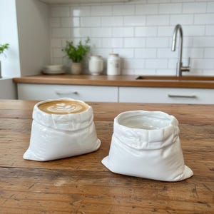 May include: Two white ceramic cups shaped like crumpled paper bags. One cup is filled with a latte featuring heart-shaped foam art, while the other cup is empty. The cups have a glossy finish and irregular, folded edges resembling a soft material.