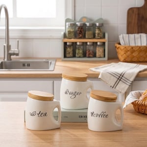 May include: Three white ceramic canisters with slanted bamboo lids and integrated handles. Each canister features black cursive text and icons: one labeled tea with a leaf icon, one labeled sugar with a whisk icon, and one labeled coffee with a bean icon.