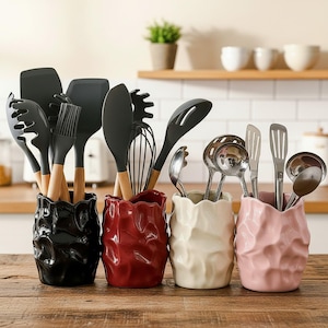 May include: Four ceramic utensil holders in black, red, cream, and light pink with a textured, crinkled surface. The holders contain various kitchen tools, including black silicone spatulas, whisks, and silver metal ladles and slotted spoons.