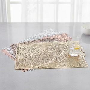 May include: A set of rectangular placemats featuring an intricate laser-cut floral and geometric pattern in metallic gold, copper, silver, and white. The mats are layered, and a clear glass filled with ice and a lemon slice sits on the top mat.
