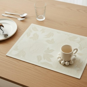 May include: A rectangular cream-colored placemat featuring a subtle, embossed floral pattern with a decorative border. A ceramic mug with a unique base made of small, spherical beads sits on the placemat, accompanied by a plate and silverware.