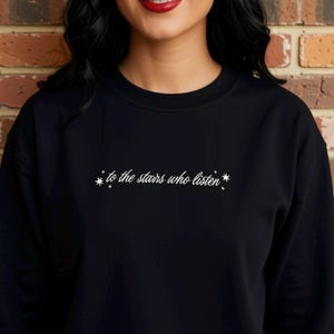 Puede incluir: Una sudadera negra de cuello redondo con texto bordado en blanco en el centro del pecho que dice "to the stars who listen". El texto está flanqueado por pequeños y delicados iconos de estrellas a ambos lados, creando un diseño limpio y sencillo.
