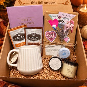 May include: A cardboard gift box containing a lavender book titled Just a Thought, a white ribbed ceramic mug, two Cocoa Classics packets, a brown paper envelope with stickers, a small jar of body cream, a candle, and a blue circular bath bomb.