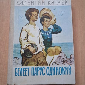Puede incluir: Un libro de tapa dura con cubierta blanca que presenta una ilustración en color de dos figuras junto al mar. El texto en la parte superior dice VALENTIN KATAEV y el texto en la parte inferior dice BELEET PARUS ODINOKIY. Las figuras visten ropa clara y oscura.