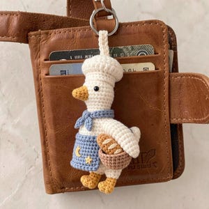 Op de afbeelding: Een gehaakte sleutelhanger van een witte eend met een koksmuts, een blauwe halsdoek en een blauw schort met gele maan- en sterborduursels. De eend houdt een klein bruin mandje met een stokbrood vast en heeft gele voeten en een gele snavel.