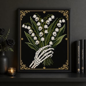 May include: A framed cross-stitch artwork on a black background featuring a skeletal hand holding a bouquet of lily of the valley flowers where each individual bell-shaped blossom is depicted as a small white skull with dark eye sockets and teeth.