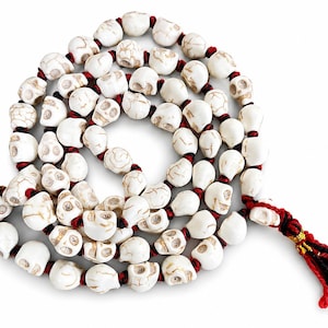 May include: A long strand of beads shaped like small skulls, crafted from white material with brown veining. The skull beads are strung together on a dark red cord, and the strand terminates in a decorative tassel made of red and black threads.