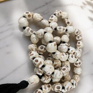 May include: A strand of beads shaped like small skulls, crafted from white material with brown veining. The skull beads are strung together on a black cord, and the strand terminates with a long, black fabric tassel at one end of the necklace.