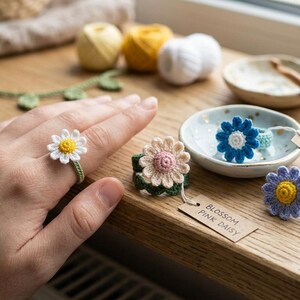 May include: A collection of handmade crochet flower rings featuring white, pink, blue, and purple petals with yellow or pink centers. Each ring has a green band and a small brown paper tag labeled with names like Blossom Pink Daisy and Azure Sky Daisy.