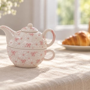 May include: A ceramic tea-for-one set featuring a teapot stacked on a teacup. The off-white surface is decorated with a repeating pattern of white daisy flowers with pink centres and small pink bows. The teapot has a curved spout and handle.