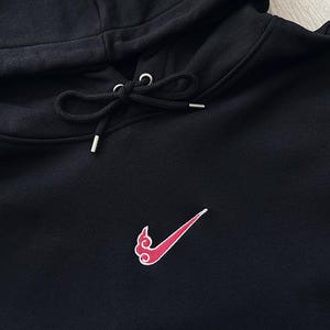 Puede incluir: Una sudadera negra con capucha y cordón ajustable con herretes metálicos. En el centro del pecho presenta un bordado de una nube roja con contorno blanco, estilizada con la forma de un símbolo de verificación diagonal o swoosh.