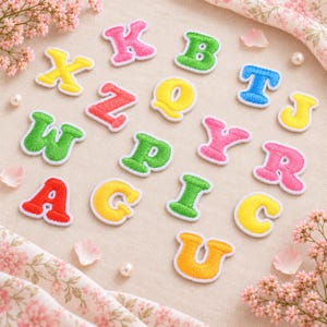 May include: A collection of embroidered alphabet letter patches in various colors including pink, green, yellow, blue, and red. Each letter features a thick white border and a textured stitched surface, arranged in a scattered pattern on a fabric surface.