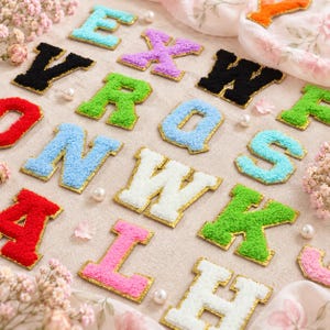 Puede incluir: Una colección de parches individuales con letras del alfabeto que presentan una superficie texturizada y difusa en varios colores, incluyendo rojo, azul, verde, rosa, blanco, morado y negro, cada uno con un borde dorado metálico sobre tela neutra.