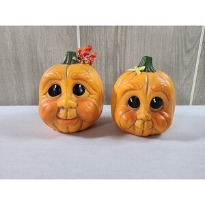 May include: Two orange pumpkin figurines with carved facial features, large black eyes, and green stems. The left pumpkin is adorned with a cluster of small red berries and dried foliage, while the right pumpkin features a small yellow ribbon bow.