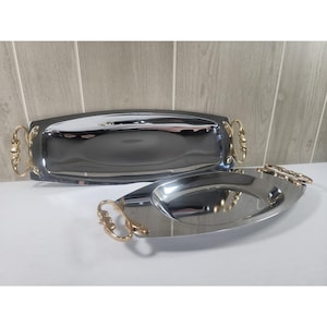 May include: Two polished silver-tone metal serving trays with reflective surfaces and ornate gold-tone metal handles on each side. The trays feature a smooth, elongated oval shape with a raised rim, showcasing a metallic finish and decorative accents.