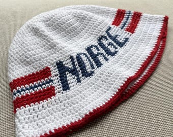 Könnte beinhalten: Eine weiße gehäkelte Mütze mit dem Wort NORGE in dunkelblauen Buchstaben, akzentuiert durch horizontale rote und blaue Streifen, die an die norwegische Flagge erinnern. Die Mütze hat einen einfarbigen roten Rand am unteren Saum und eine Textur aus Garn.