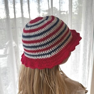 May include: A crochet bucket hat featuring horizontal stripes in red, white, and dark blue. The hat has a rounded crown and a wide, scalloped red brim. The construction consists of dense, textured yarn stitches throughout the entire design.