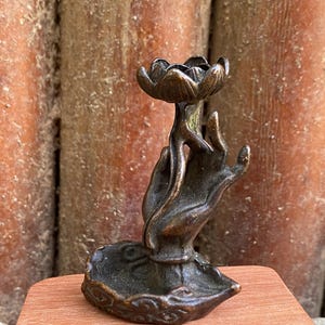 May include: A bronze-toned metal incense holder featuring a sculpted hand rising from a circular base, holding a lotus flower-shaped cup at the top. The object has a dark, weathered finish with visible texture and intricate detailing on the hand and base.