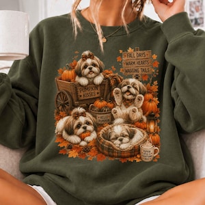 Pode incluir: Uma camisola de gola redonda verde-azeitona com uma impressão gráfica de quatro cães Shih Tzu rodeados por abóboras, folhas de outono e uma placa de madeira com a inscrição FALL DAYS WARM HEARTS WAGGING TAILS, PUMPKIN KISSES e HELLO PUMPKIN.