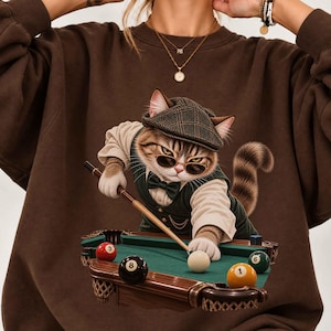 Peut inclure: Un sweat-shirt à col rond marron foncé orné d'un motif représentant un chat tigré portant une casquette plate à carreaux, des lunettes de soleil, un gilet et un nœud papillon. Le chat est devant un billard vert, tenant une queue de billard.