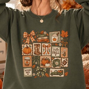 Könnte beinhalten: Ein olivgrüner Rundhals-Pullover mit einem zentralen Grafikdruck verschiedener herbstlicher Motive, darunter Kürbisse, Cowboystiefel, Schleifen, eine Kaffeetasse, ein Football, ein Truck und Bücher mit der Aufschrift "COZY FALL VIBES" und "HELLO FALL".