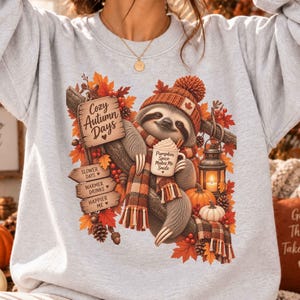 Könnte beinhalten: Ein hellgraues Sweatshirt mit einer Grafik eines Faultiers, das eine Strickmütze und einen Schal trägt und eine Tasse mit der Aufschrift Pumpkin Spice Makes Me Smile hält. Das Design zeigt Herbstblätter, eine Laterne, Kürbisse und Holzschilder.