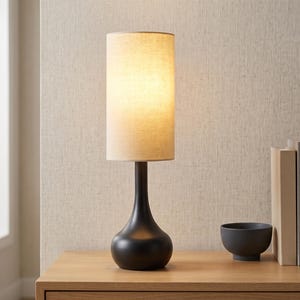 May include: A table lamp with a matte black teardrop-shaped base and a tall, cylindrical beige fabric shade. The lamp emits a warm, soft glow from within the shade, which is positioned upright on a flat surface next to a small bowl and books.