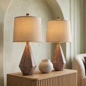 May include: Two table lamps with faceted, dark wood-toned bases and cylindrical, light beige fabric shades. Between the lamps sits a small, round, off-white ceramic vase with a textured finish. The lamps feature a simple, geometric design.